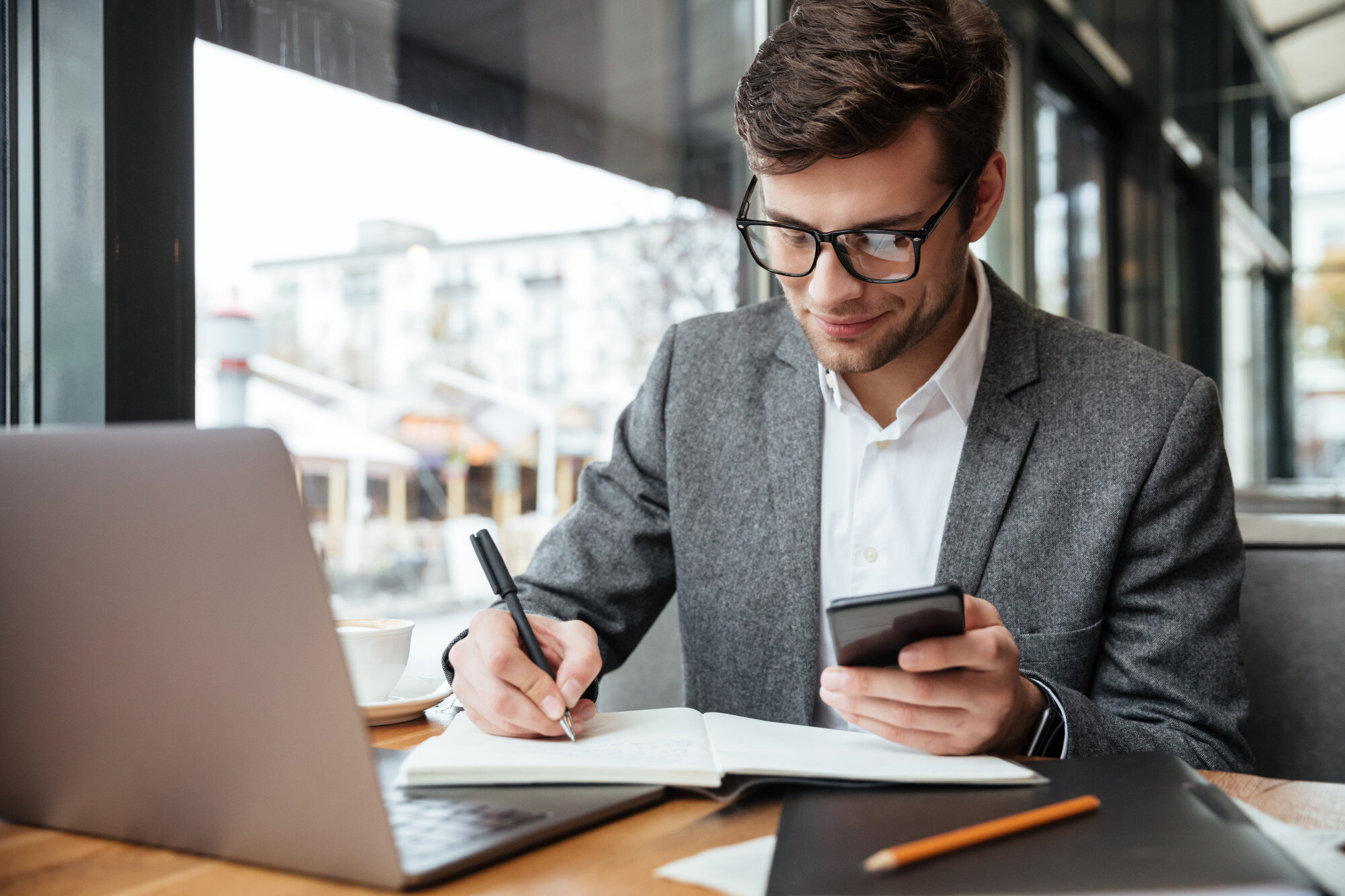 smiling-businessman-eyeglasses-sitting-by-table-cafe-with-laptop-computer-while-using-smartphone-writing-something